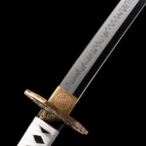 Puede incluir: Primer plano de una katana japonesa. La hoja plateada presenta un patr&oacute;n detallado, y la guarda tiene detalles dorados y negros. El mango est&aacute; envuelto en tela blanca. La espada se encuentra sobre un fondo negro.