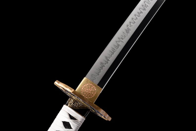 Puede incluir: Primer plano de una katana japonesa. La hoja plateada presenta un patr&oacute;n detallado, y la guarda tiene detalles dorados y negros. El mango est&aacute; envuelto en tela blanca. La espada se encuentra sobre un fondo negro.