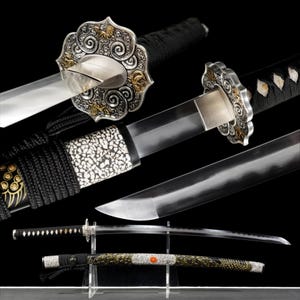 Puede incluir: Una katana con empuñadura envuelta en cordón negro, una guarda plateada con intrincados grabados y detalles dorados, y una hoja de acero pulido. La vaina es negra con incrustaciones blancas texturizadas y un detalle circular naranja.