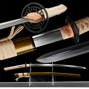 May include: A Japanese katana sword with a curved, polished steel blade. The hilt is wrapped in beige fabric, and the guard is black with a decorative design. The scabbard is gold with a matching beige fabric wrap.