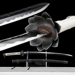 May include: A Japanese katana sword with a black and silver blade, a white handle, and a decorative floral guard. The sword is displayed with its black scabbard on a clear stand against a black background.