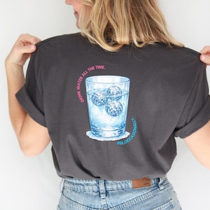 Può includere: T-shirt grigio scuro con grafica di un bicchiere d'acqua con tre cubetti di ghiaccio a sfera da discoteca. Il testo "DRINK WATER ALL THE TIME" e "PEE.OCCASIONALLY." è stampato in rosa e blu.