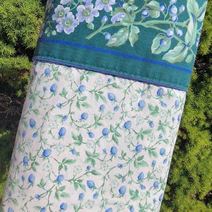 May include: A pillowcase with a floral design. The top portion is a dark teal with a floral pattern of light blue flowers and green leaves. The bottom portion is white with a pattern of blue berries and green leaves.