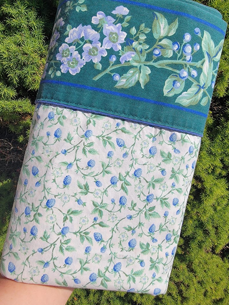 May include: A pillowcase with a floral design. The top portion is a dark teal with a floral pattern of light blue flowers and green leaves. The bottom portion is white with a pattern of blue berries and green leaves.