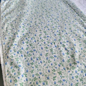 May include: A floral patterned fabric featuring a repeating design of blue berries and green leaves on a white background. The fabric has a border of blue and green floral patterns. The fabric is likely used for quilting or other sewing projects.