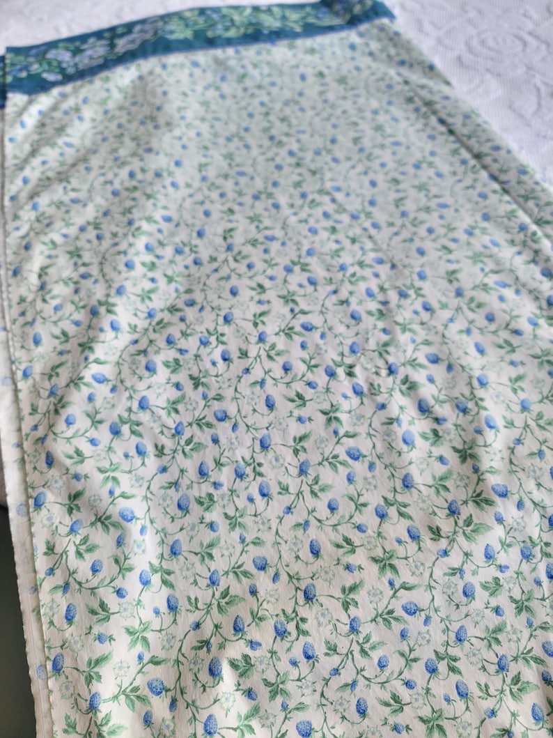 May include: A floral patterned fabric featuring a repeating design of blue berries and green leaves on a white background. The fabric has a border of blue and green floral patterns. The fabric is likely used for quilting or other sewing projects.