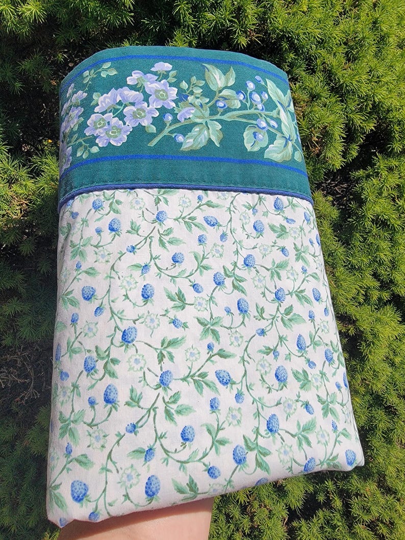 May include: A pillowcase with a floral design. The top portion is teal with a floral pattern in shades of blue and green. The bottom portion is white with a pattern of blue berries and green leaves.