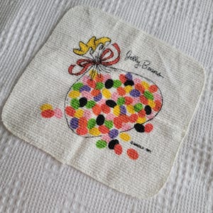 K&ouml;nnte beinhalten: Wei&szlig;es quadratisches Handtuch mit einer farbenfrohen Illustration eines Beutels mit Geleebohnen. Der Beutel ist mit einer roten Schleife und einem gelben Band gebunden. Die Worte "Jelly Beans" sind auf dem Handtuch geschrieben.