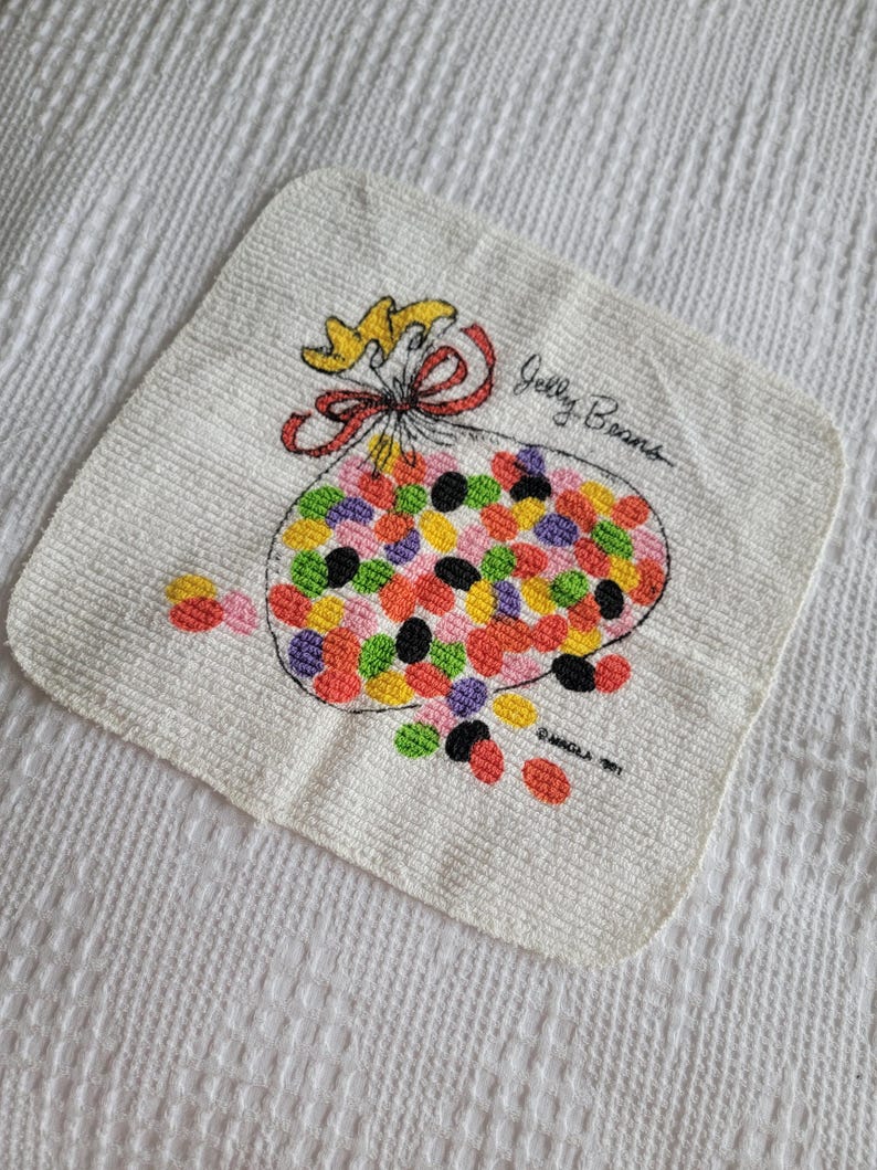 K&ouml;nnte beinhalten: Wei&szlig;es quadratisches Handtuch mit einer farbenfrohen Illustration eines Beutels mit Geleebohnen. Der Beutel ist mit einer roten Schleife und einem gelben Band gebunden. Die Worte "Jelly Beans" sind auf dem Handtuch geschrieben.