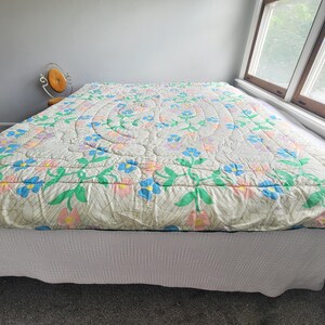 May include: A floral quilt with a cream background, featuring blue, pink, and green flower patterns. The quilt is placed on a bed with a white bed skirt. The room has a window and a small chair.