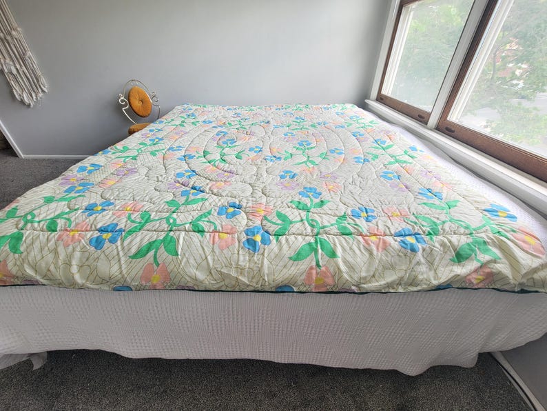 May include: A floral quilt with a cream background, featuring blue, pink, and green flower patterns. The quilt is placed on a bed with a white bed skirt. The room has a window and a small chair.