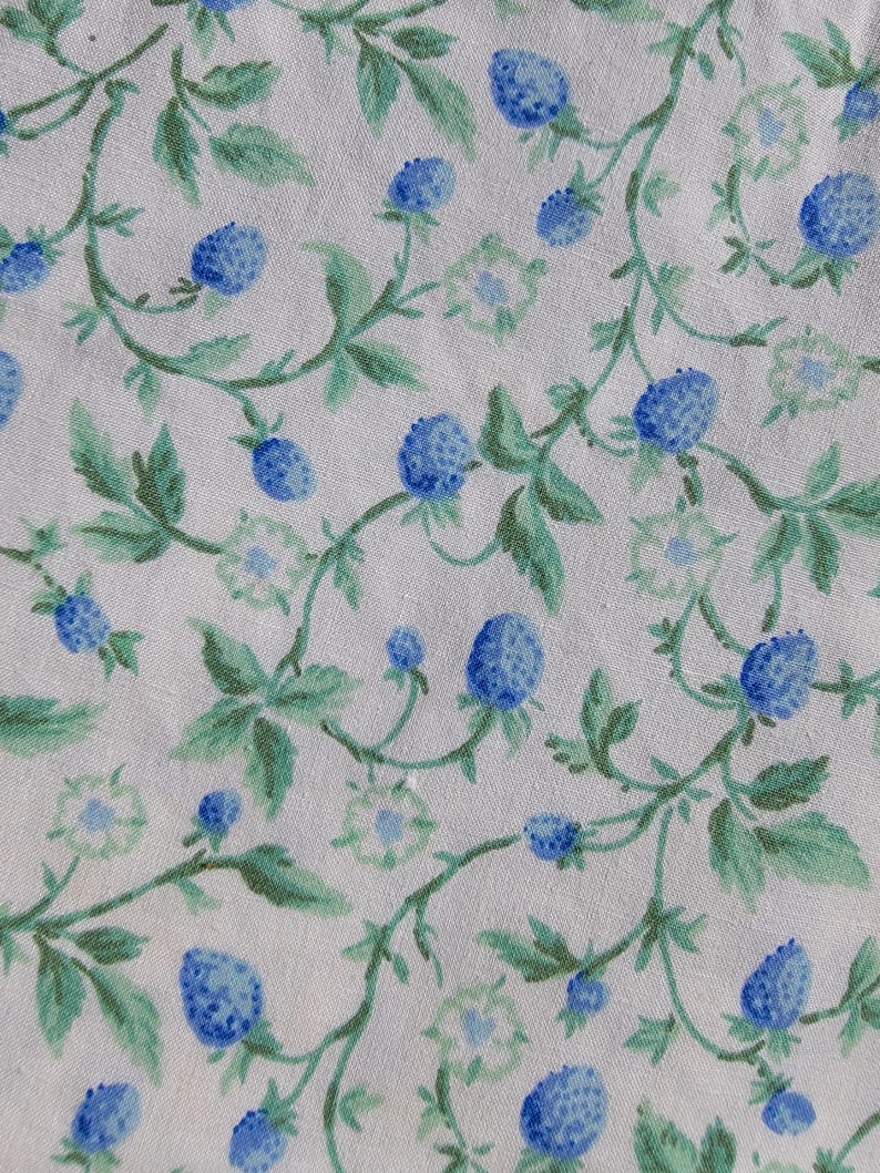 May include: A fabric with a repeating pattern of blue berries and green leaves on a white background. The berries are a vibrant blue, and the leaves are a mix of light and dark green. The design has a watercolor effect.