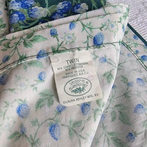 May include: Twin-sized floral bed sheet set with a white background and a pattern of blue berries and green leaves. The label reads "Laura Ashley" and "Made in U.S.A."