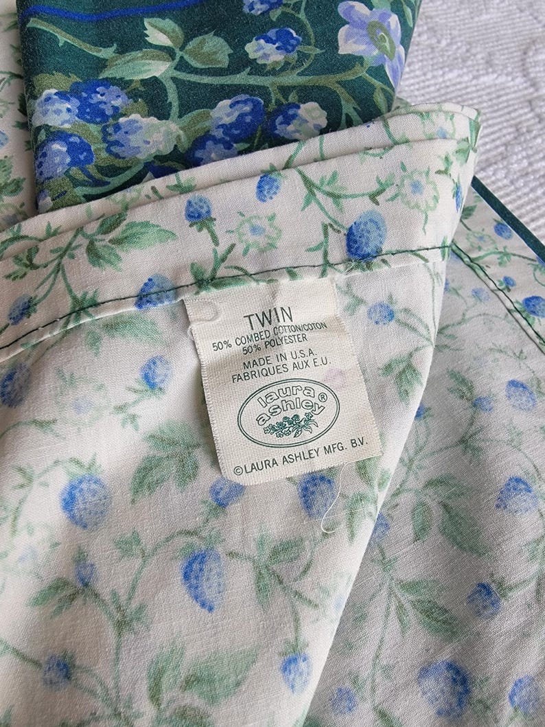 May include: Twin-sized floral bed sheet set with a white background and a pattern of blue berries and green leaves. The label reads "Laura Ashley" and "Made in U.S.A."