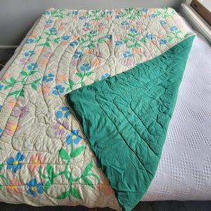 May include: A floral-patterned quilt with a cream background, featuring blue, purple, and pink flowers with green stems. The quilt is partially folded back, revealing a solid teal green underside. The quilt is on a bed.