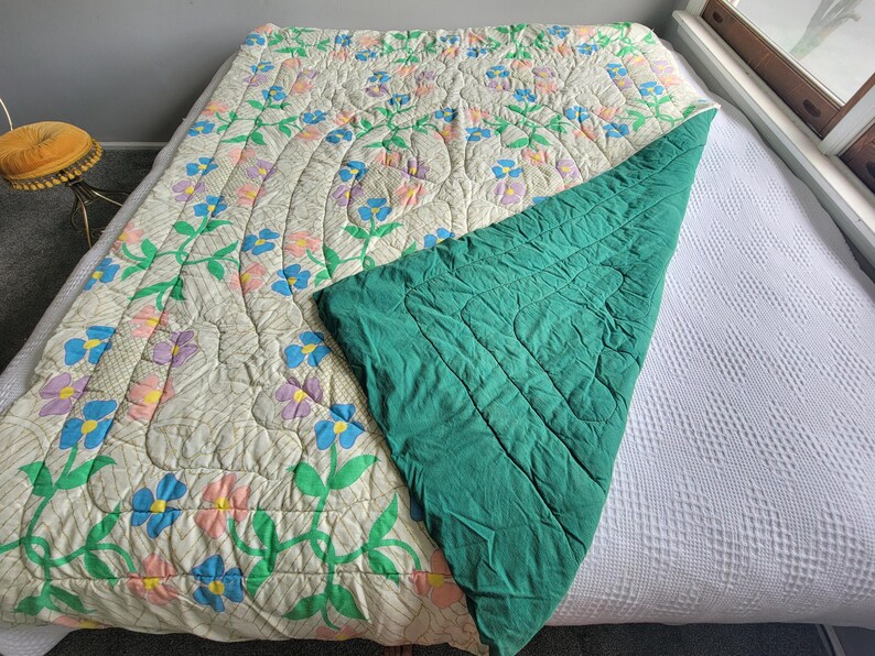 May include: A floral-patterned quilt with a cream background, featuring blue, purple, and pink flowers with green stems. The quilt is partially folded back, revealing a solid teal green underside. The quilt is on a bed.