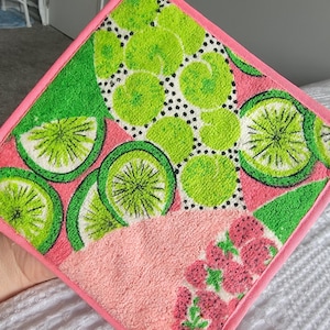 May include: A square, pink and green pot holder with a loop for hanging. The pot holder features a lime and floral design. The border and hanging loop are a solid pink color. The lime design includes green and white details.