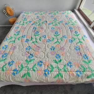 May include: A cream-colored quilt with a floral pattern in blue, pink, and purple. The quilt is spread over a bed, with a repeating design of flowers and leaves. The quilt's dimensions are not visible.