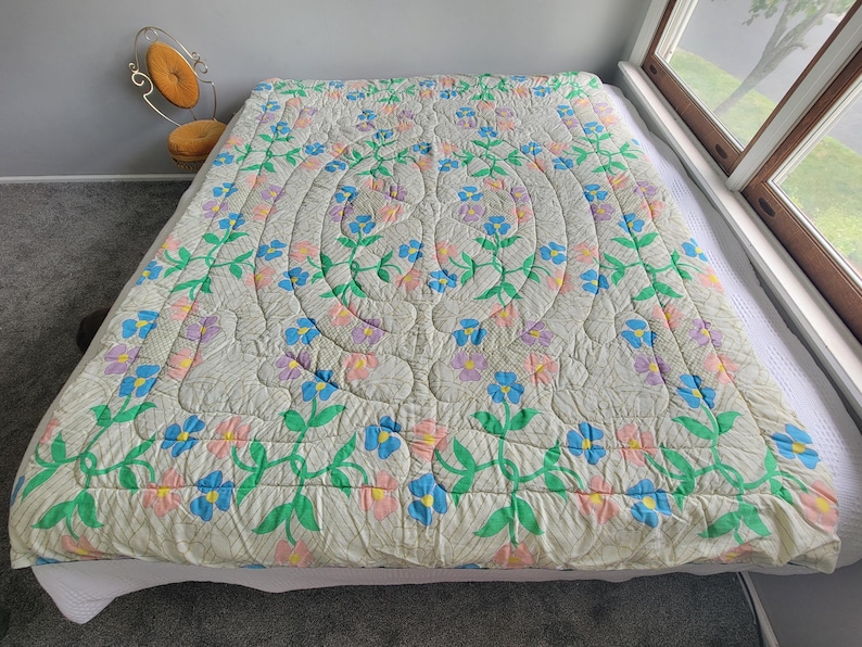 May include: A cream-colored quilt with a floral pattern in blue, pink, and purple. The quilt is spread over a bed, with a repeating design of flowers and leaves. The quilt's dimensions are not visible.