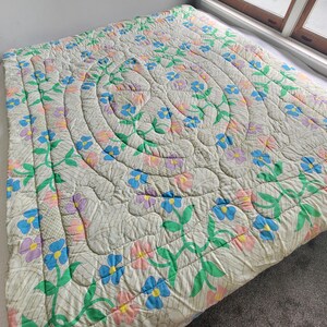 May include: A floral quilt with a cream background, featuring a central spiral design and repeating floral patterns in blue, pink, and purple. Green leaves and stems accent the flowers. The quilt is on a bed.