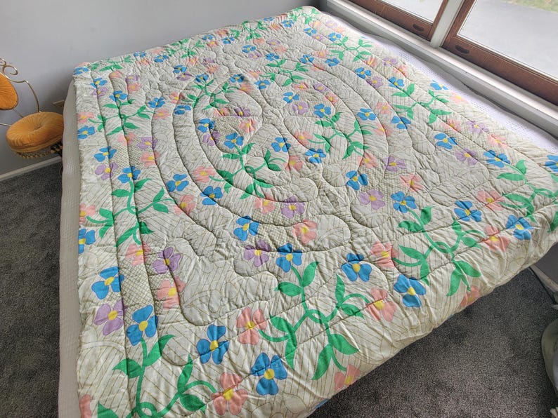 May include: A floral quilt with a cream background, featuring a central spiral design and repeating floral patterns in blue, pink, and purple. Green leaves and stems accent the flowers. The quilt is on a bed.
