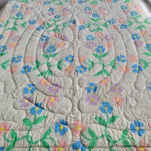 May include: A quilted bedspread with a floral design. The off-white fabric is patterned with a repeating grid. The quilt features a central design of concentric circles with stylized flowers in blue, pink, and purple, with green stems and leaves.