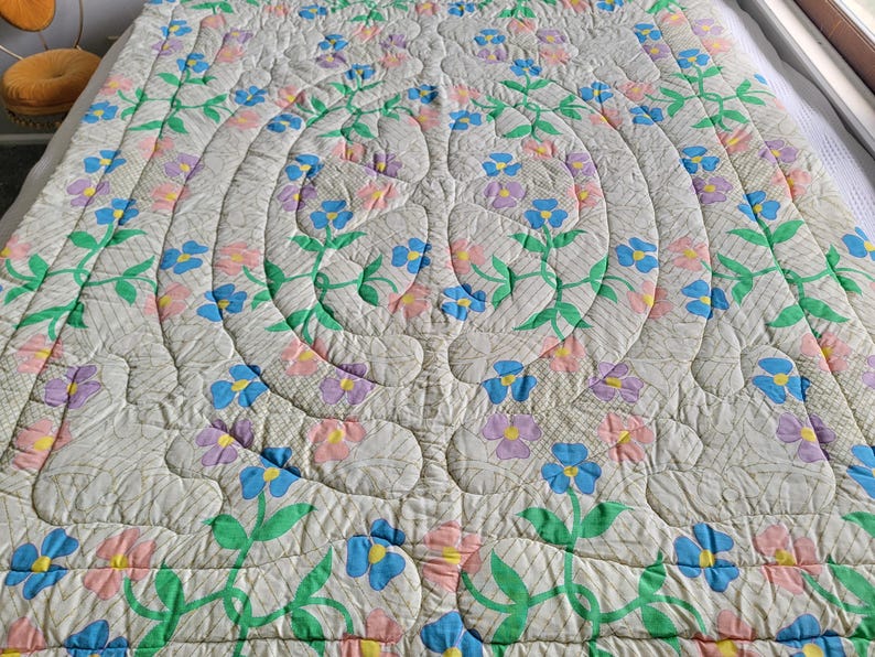 May include: A quilted bedspread with a floral design. The off-white fabric is patterned with a repeating grid. The quilt features a central design of concentric circles with stylized flowers in blue, pink, and purple, with green stems and leaves.