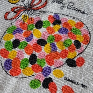 K&ouml;nnte beinhalten: Wei&szlig;es Handtuch mit einem gestickten Design eines Beutels mit bunten Geleebohnen. Der Beutel ist mit einer roten Schleife und gelben Akzenten gebunden. Die Worte "Jelly Beans" sind &uuml;ber dem Beutel gestickt. Das Handtuch ist strukturiert.