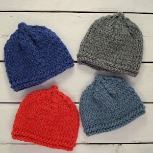 May include: Four knitted hats in different colours: blue, grey, red, and light blue. The hats are arranged in a square pattern on a white wooden surface.