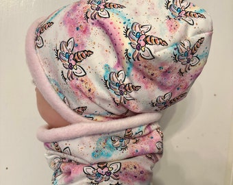 May include: A white knit hat and neck warmer set with a unicorn print. The hat has a pink fleece lining. The print features colorful unicorns with glitter and a rainbow background.