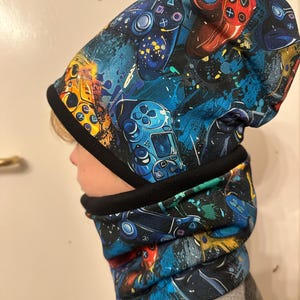 Könnte beinhalten: Eine schwarze Mütze und ein Nackentuch mit einem bunten Muster aus Videospielcontrollern und blauen, gelben und roten Farbflecken.