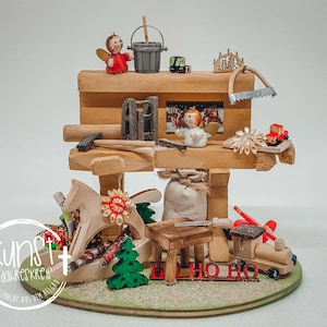 Könnte beinhalten: Eine hölzerne Weihnachtswerkstatt-Szene mit Miniaturwerkzeugen, Spielzeug und Figuren. Die Szene umfasst einen kleinen Engel, einen Eimer, einen Hammer, eine Säge, ein Schaukelpferd und einen Zug. Die Basis ist ein grüner Kreis, und das Gesamtdesign ist rustikal und festlich.