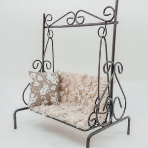 Puede incluir: Columpio de metal en miniatura con un diseño decorativo en espiral. El columpio presenta un asiento beige y esponjoso y una pequeña almohada con un estampado floral blanco. El columpio está diseñado para fines decorativos.
