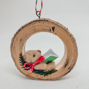 Può includere: Un ornamento natalizio con un piccolo orsacchiotto che legge un libro, all'interno di una ghirlanda di legno. L'orso ha un fiocco rosso ed è circondato da ramoscelli verdi. La ghirlanda è sospesa da una corda rossa, verde e bianca.