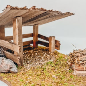 Könnte beinhalten: Ein Miniatur-Holzstall mit rustikalem Design. Die Struktur verfügt über einen Lattenzaun, ein Holzdach und eine mit Moos und Heu bedeckte Basis. Ein Holzstapel und ein Stein sind ebenfalls vorhanden.