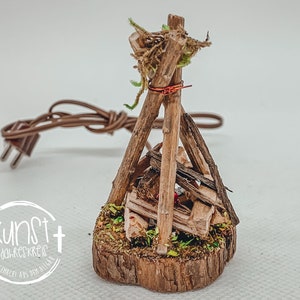 Pode incluir: Lâmpada de fogueira em miniatura feita de madeira, musgo e galhos. A lâmpada apresenta uma pequena base de madeira e um cabo elétrico marrom com um plugue. A fogueira é construída com pequenos troncos e galhos de madeira e é coberta com musgo.