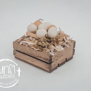 Puede incluir: Una pequeña caja de madera llena de paja y huevos falsos. La caja es marrón y tiene un aspecto rústico. Los huevos son blancos y de color crema.