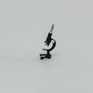 Può includere: Un microscopio nero in miniatura con dettagli argentati. Il piccolo strumento scientifico è posto su uno sfondo bianco. Il microscopio ha una base, un tavolino, un braccio curvo e un oculare.