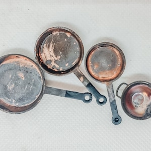 Può includere: Quattro padelle in rame vintage con manici neri. Le padelle sono di diverse dimensioni e hanno un aspetto usurato e rustico.