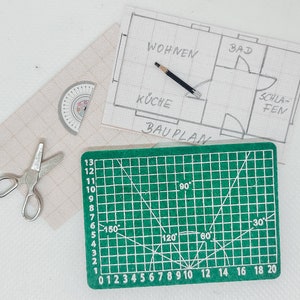 Könnte beinhalten: Eine grüne Schneidematte mit Markierungen zum Messen von Winkeln und einem Lineal. Die Matte liegt auf einer weißen Oberfläche mit einem Bleistift, einer Schere und einem Bauplan für ein Haus.