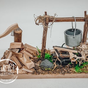 Könnte beinhalten: Eine Miniatur-Rustikalszene mit einer Holzaxt, einem Stapel Brennholz und einem kleinen Metalleimer, der an einem Holzrahmen hängt. Die Basis ist mit Moos und kleinen Steinen bedeckt. Der Text "kunst jahreskreis landgemacht aus dem allgäu" ist enthalten.