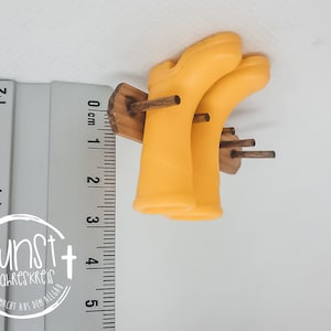 Könnte beinhalten: Ein Paar gelbe Gummistiefel hängen an einem Holzgestell. Das Gestell hat zwei Holzstift und ist an einer Holzbasis befestigt. Die Stiefel sind etwa 12,5 cm hoch.