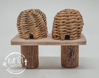 Può includere: Due piccole case a forma di alveare, intrecciate, sono posizionate su una piattaforma di legno con due gambe di legno. Gli alveari sono realizzati con materiali naturali e hanno un colore marrone chiaro.