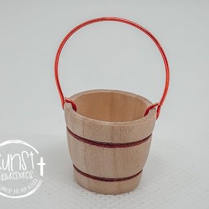 Puede incluir: Un peque&ntilde;o cubo de madera con un asa de alambre rojo. El cubo tiene un acabado de madera natural y rayas rojas alrededor de la parte superior.