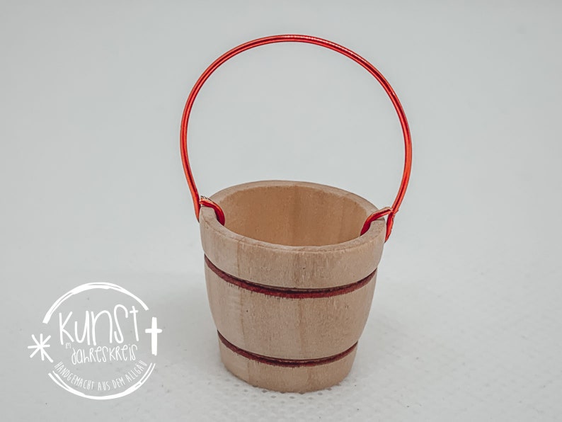 Puede incluir: Un peque&ntilde;o cubo de madera con un asa de alambre rojo. El cubo tiene un acabado de madera natural y rayas rojas alrededor de la parte superior.