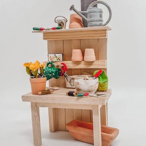 Könnte beinhalten: Miniatur-Gartentisch aus Holz mit Gartengeräten, darunter eine Gießkanne, Töpfe und ein kleiner Blumentopf mit gelben Blumen. Die Werkbank hat ein Regal mit kleinen Terrakottatöpfen und ein kleines rechteckiges Schild.