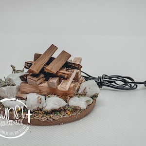 Könnte beinhalten: Ein Miniatur-Holzfeuer mit einem schwarzen Kabel und Stecker, umgeben von weißen Steinen und grünem Laub. Die Basis ist braun und trägt den Text "Kunst + dahinterkreis handgemacht aus dem allgäu".
