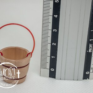 Puede incluir: Un peque&ntilde;o cubo de madera con un asa de alambre rojo. El cubo mide aproximadamente 5 cm de alto y tiene un di&aacute;metro de unos 4 cm. El cubo est&aacute; hecho de madera natural y tiene un color marr&oacute;n claro.