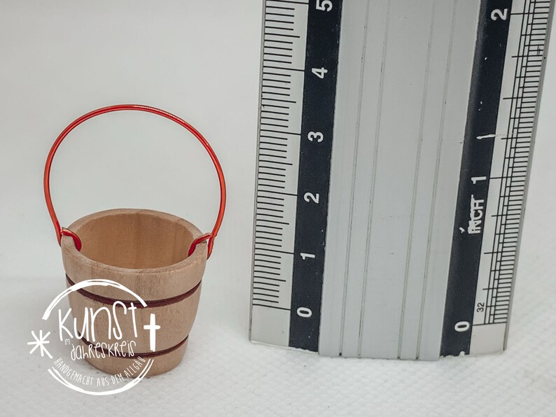 Puede incluir: Un peque&ntilde;o cubo de madera con un asa de alambre rojo. El cubo mide aproximadamente 5 cm de alto y tiene un di&aacute;metro de unos 4 cm. El cubo est&aacute; hecho de madera natural y tiene un color marr&oacute;n claro.