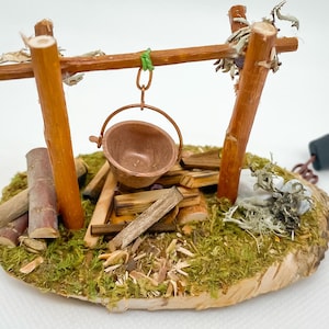Op de afbeelding: Miniatuur kampvuurscène met een koperen pot die boven een stapel hout hangt. De scène staat op een houten basis bedekt met groen mos en bevat kleine stammen en twijgen. Een zwarte stekker is bevestigd.